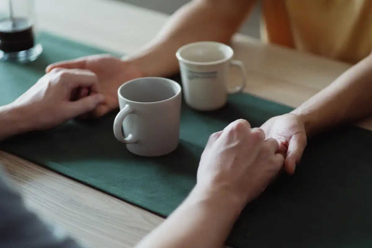 Două mâini care se țin peste o masă cu cești de cafea, semn de sprijin și apropiere