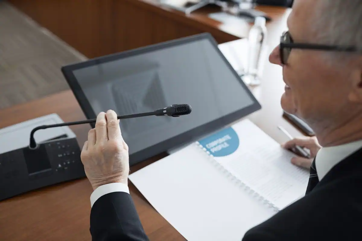 Bărbat care vorbește calm la microfon în timpul unei ședințe cu documente pe masă