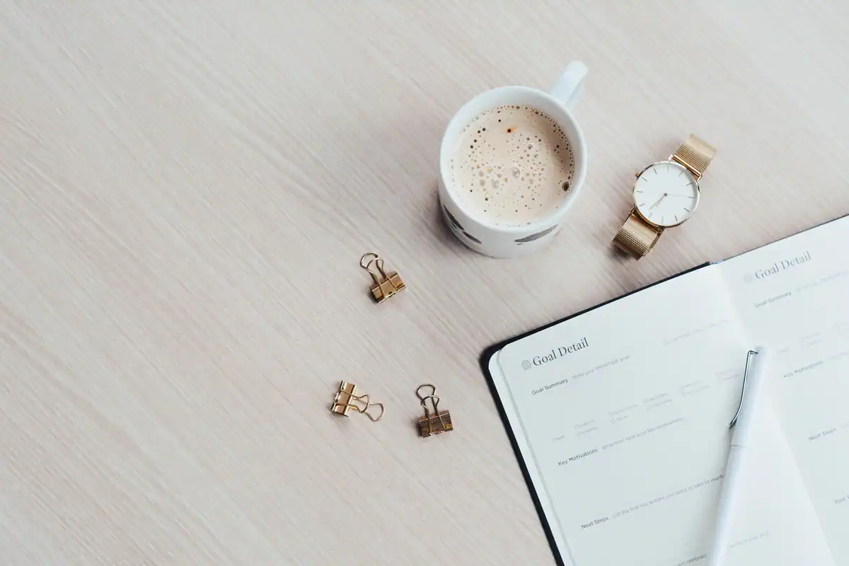 Birou minimalist cu o cană de cafea, un carnet, un pix și un ceas de mână