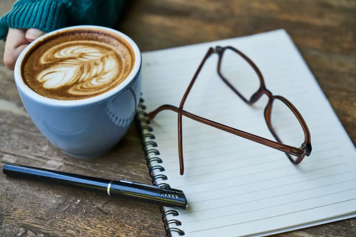 Colț de lucru cu o cafea cu spumă, un carnet și ochelari, loc pentru gânduri